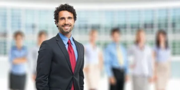 A man in corporate attire is smiling at the camera. On his blurred background are fellow executives/employees dressed in various corporate attire.